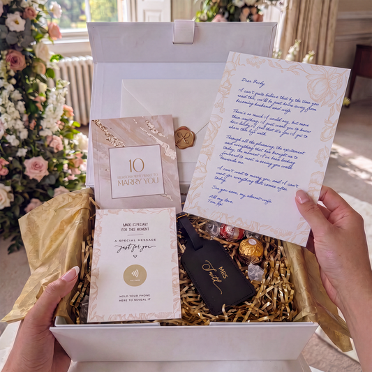A gift box opened by two hands, containing a handwritten letter held up by a hand, a '10 Years Reasons Why I want to Marry You' booklet, a 'Special Message Just for You' card, gold shredded paper, chocolates, and a black velvet box with gold lettering, set against a blurred background of pink roses.