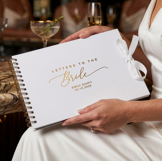 A white spiral-bound wedding guest book with gold foil text reading 'Letters to the Bride' and a white ribbon bow, held by a woman in a white dress in a dimly lit bar setting.